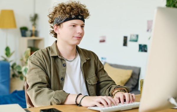 Youthful guy in casualwear looking through information on screen of desktop computer and typing on keyboard while sitting by desk